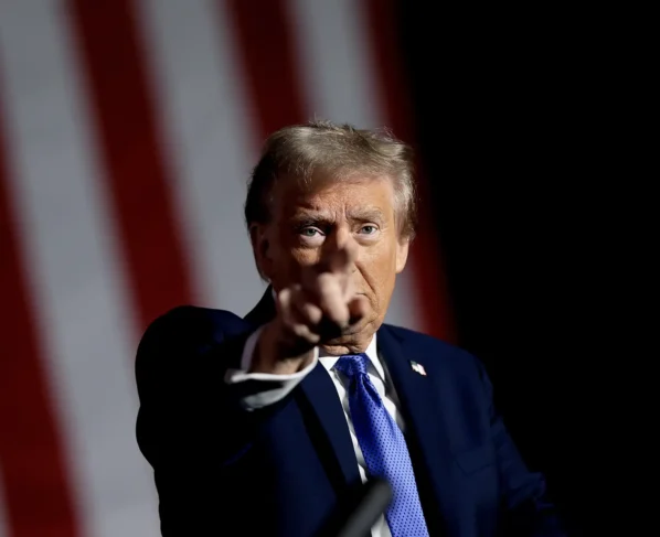 Retrato do ex-presidente dos EUA, Donald Trump, um homem idoso em terno formal azul escuro aponta enfaticamente para a câmera com expressão séria, fundo sugere uma ocasião oficial nos Estados Unidos.